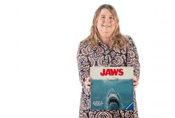 TCU film professor Kim Owczarski holds a 'Jaws'-themed board game while standing against a white backdrop.