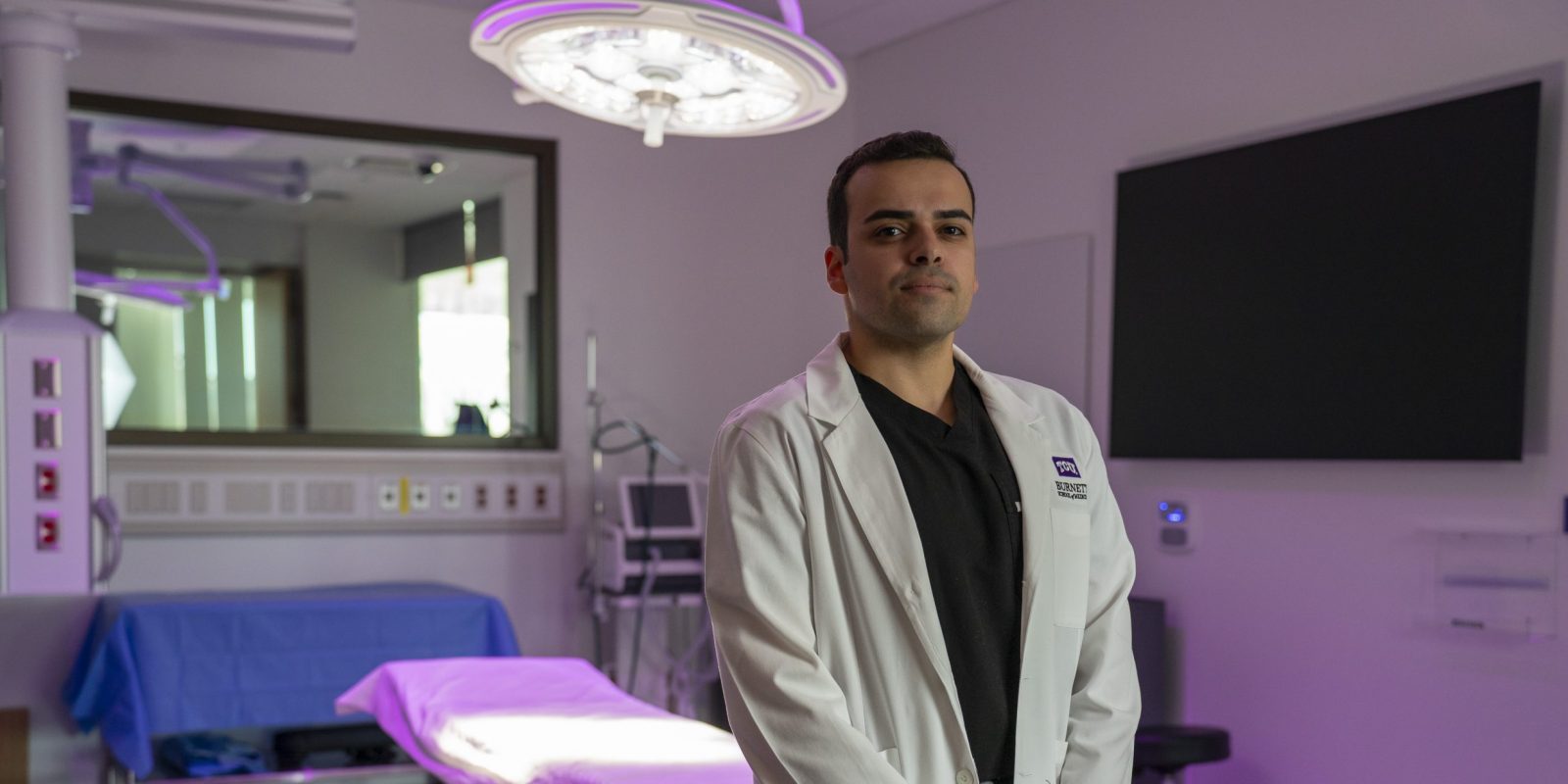 Photograph of recent MD graduate Ethan Vieira wearing a white coat, black shirt and black pants, standing in a purple-lit medical room with surgical lights, a transparent window and a large, powered-off screen.