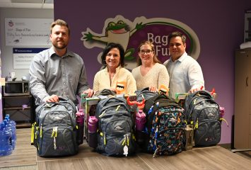 Photograph of four people standing behind a boardroom table filled with stuffed backpacks, with the 'Bags of Fun' logo on a purple wall in the background.
