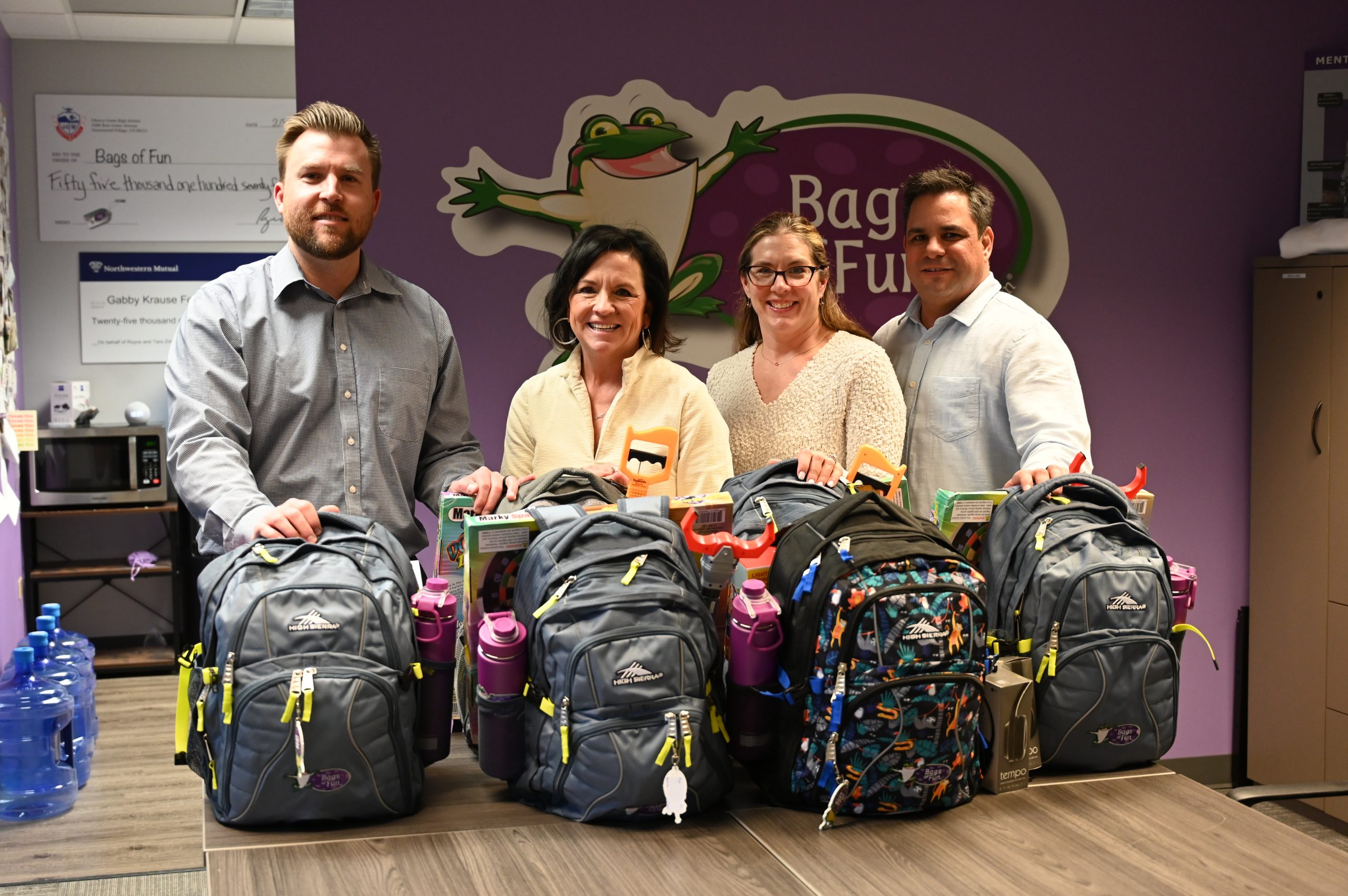 Photograph of four people standing behind a boardroom table filled with stuffed backpacks, with the 'Bags of Fun' logo on a purple wall in the background.