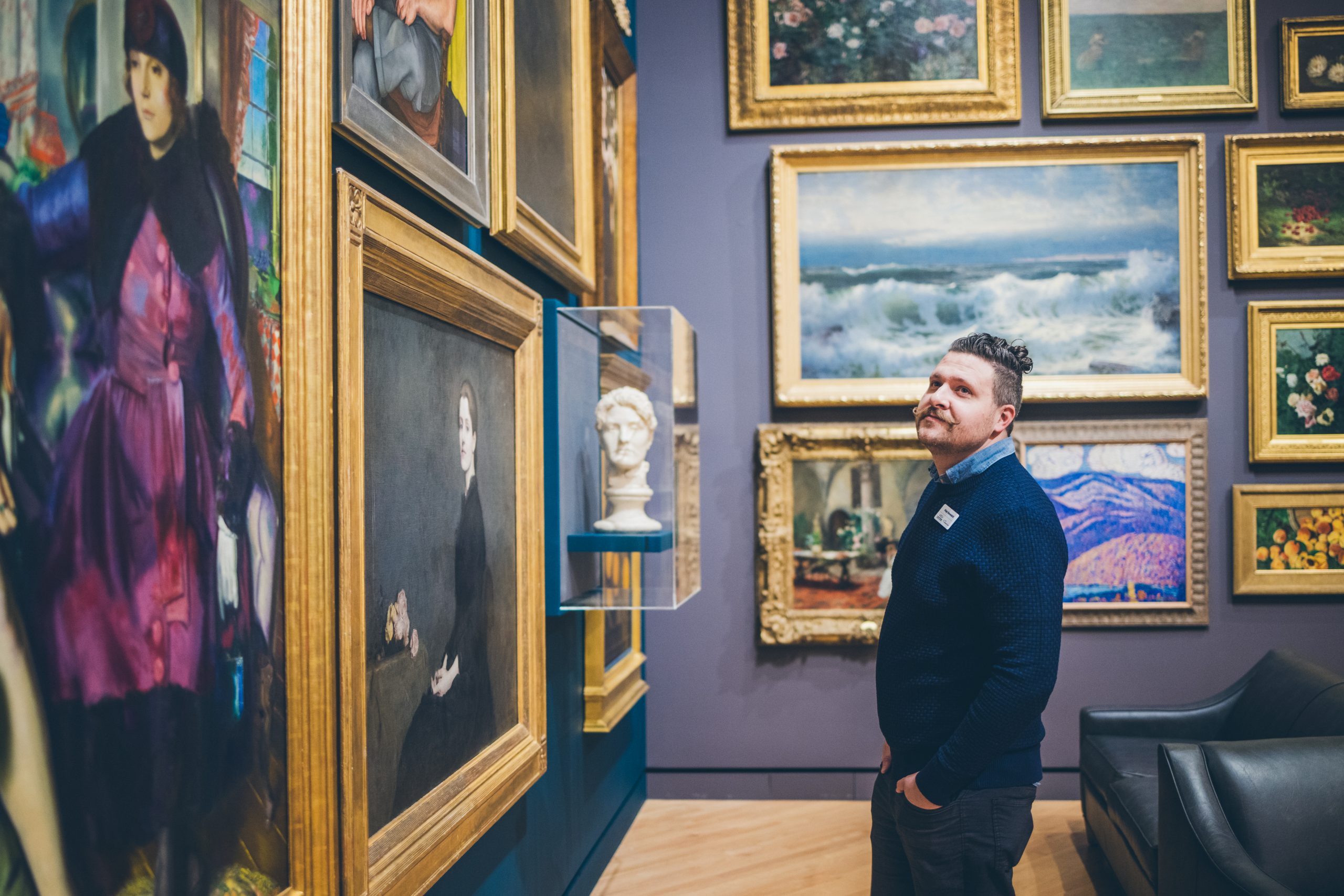 Photograph of art curator Alejo Benedetti standing with hands in his pockets in a grayish-purple-walled gallery, looking at artwork displayed on the walls.