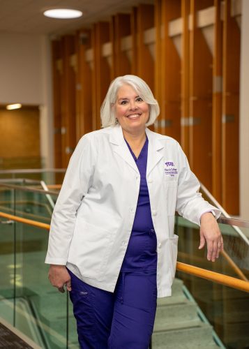 Maura Lindenfeld, Texas Christian University assistant professor of nursing, dressed in a white coat and purple scrubs, leans against a railing.