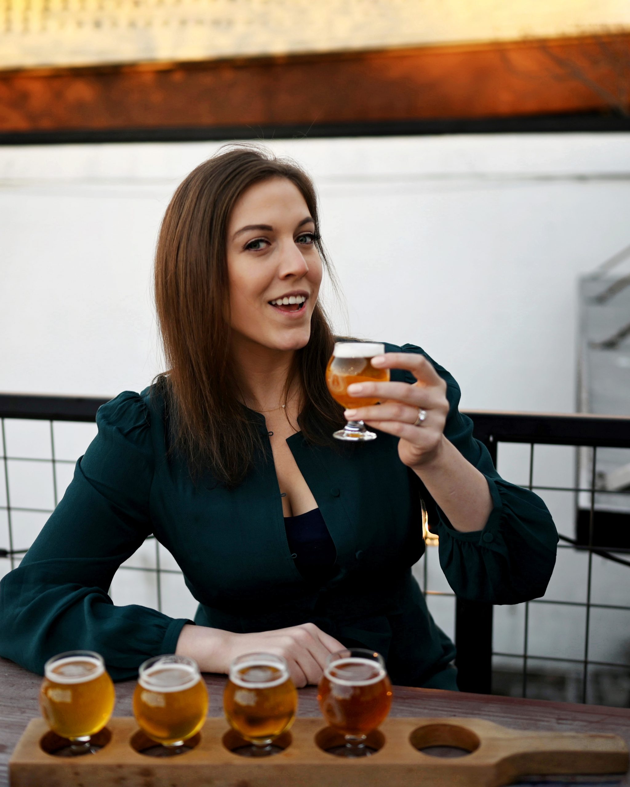 Mandy Naglich smiling while holding a sample glass of beer, seated in an outdoor porch-like setting.