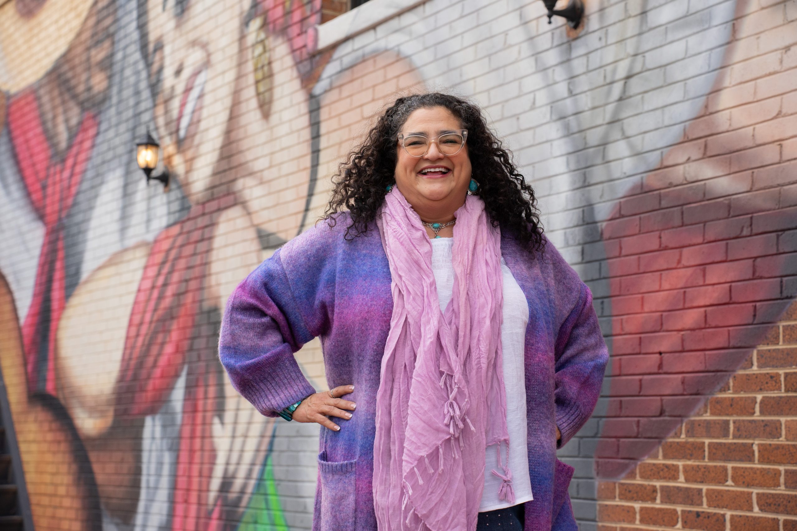 Marcela Fuentes smiles with her hands on her hips, wearing a pink scarf, long purple garment and white shirt. She stands in front of a mural depicting two people dancing in red and white clothing.