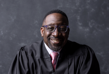 Portrait of Maryland District Court Judge Zuberi Williams, seated and smiling while wearing a black robe over a dress shirt and tie, against a gray background.