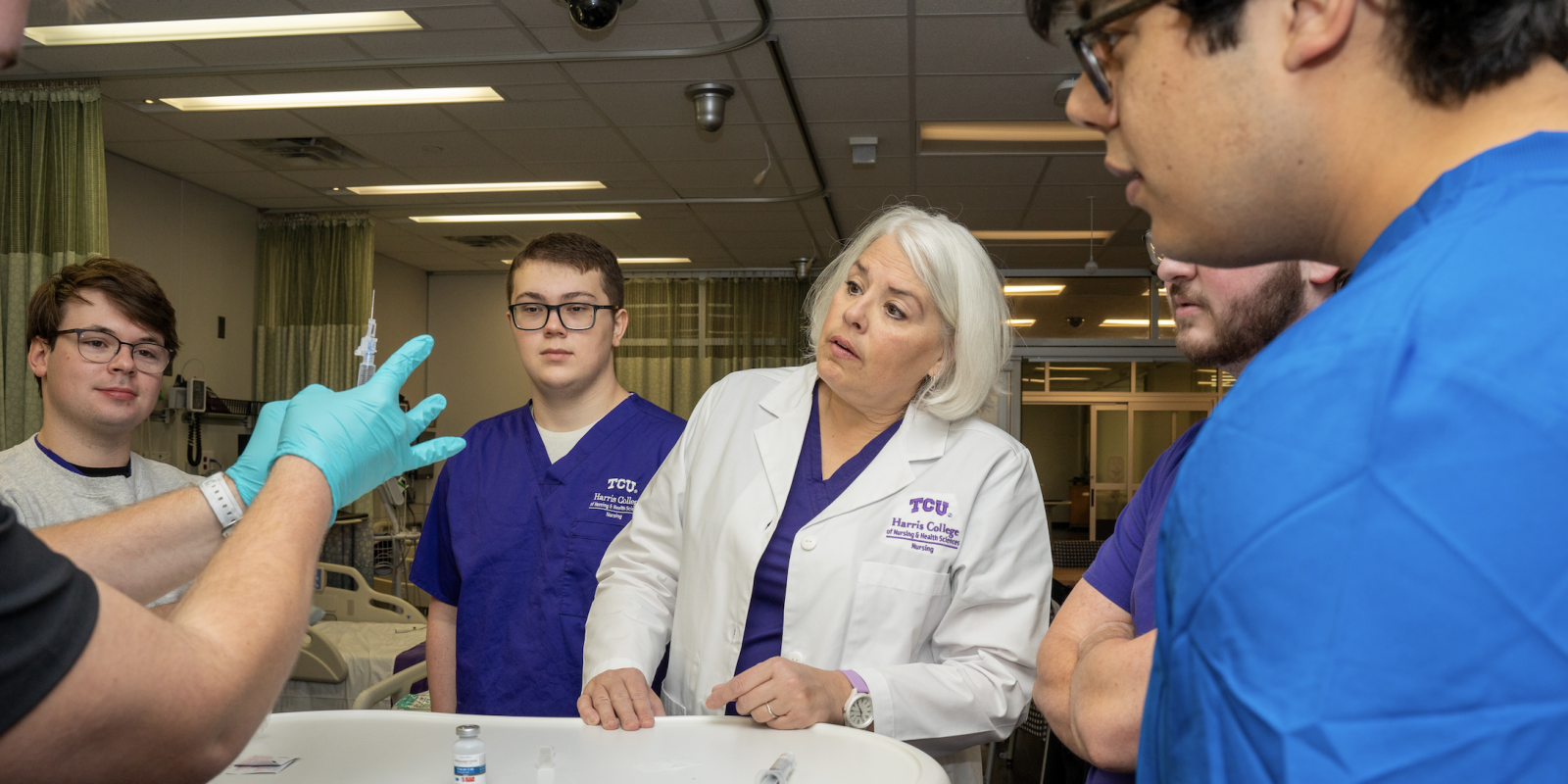Maura Lindenfeld stands with three students in scrubs, observing a fourth student in scrubs and light blue gloves who is preparing a syringe.