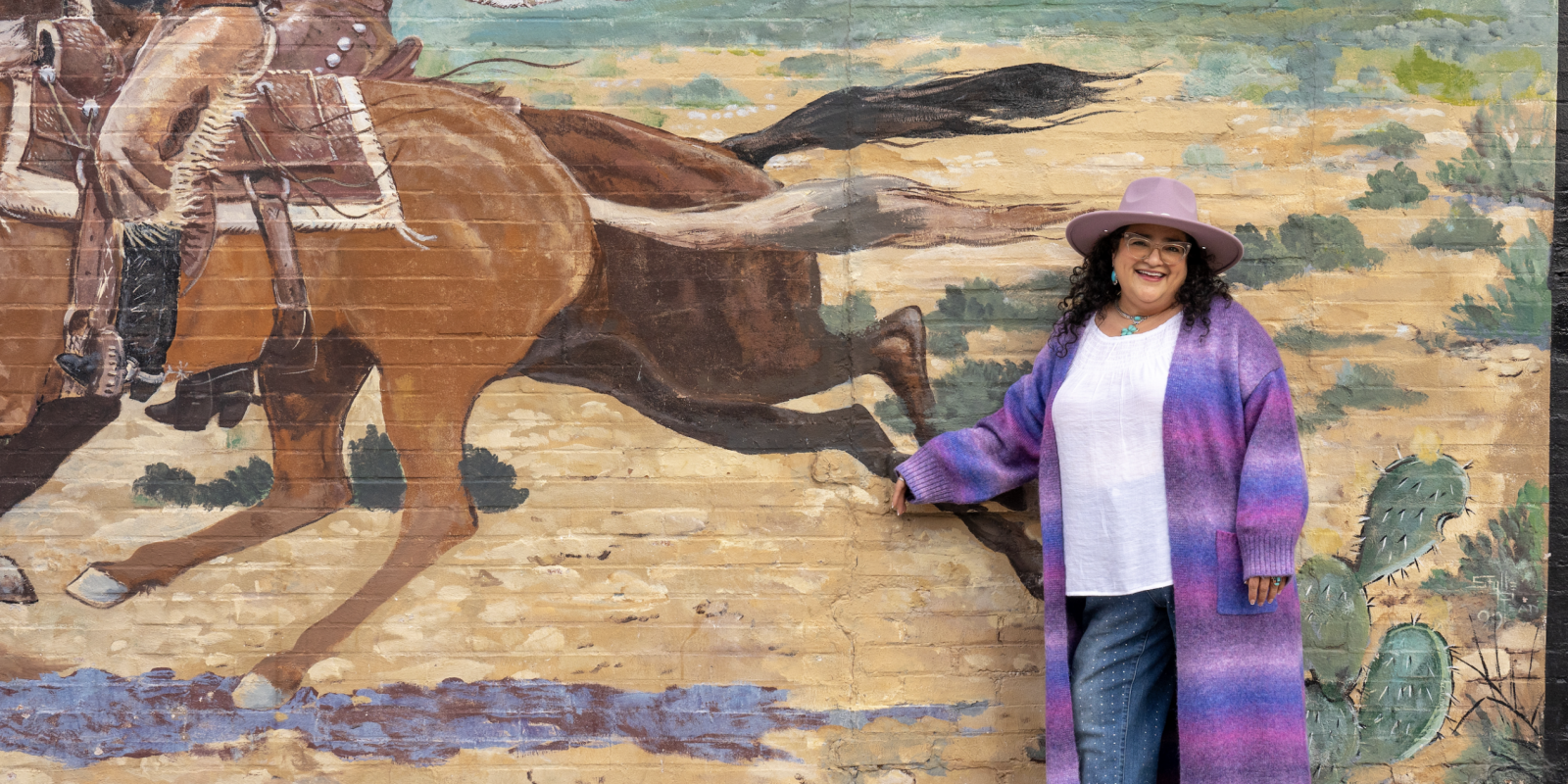 Photograph of Marcela Fuentes, author and assistant professor of English - creative writing (prose), smiling as she stands in front of a mural of a galloping brown horse. She wears a long, tri-colored purple garment over a white shirt and jeans.