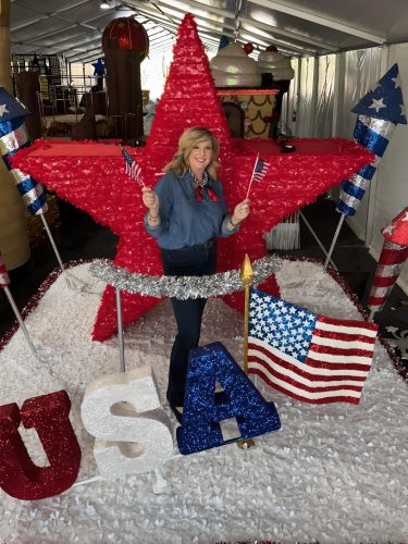 Photograph of TCU alumna Lezlie Barnson-DeNardin smiling and holding mini U.S. flags in both hands as she stands on a “USA”-themed Fourth of July parade float, looking toward the camera.