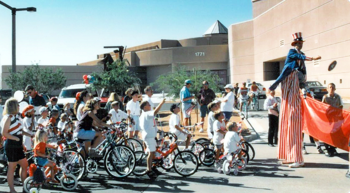 Photograph of a Fourth of July parade scene featuring a person on stilts dressed in red, white and blue standing in the street as children and adults on decorated bicycles look on.