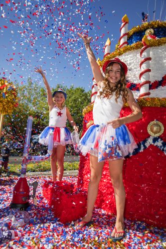 Two children in red, white and blue outfits throw confetti on a Fourth of July parade float during a sunny day.