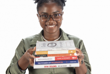 Photograph of TCU student Charity Ketu smiling at the camera while holding a stack of books. She stands against a white background, wearing black-framed glasses, a gray-green dress shirt and a black skirt.