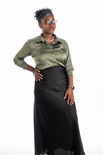 Photograph of TCU student Charity Ketu standing against a white background, wearing black-framed glasses, a gray-green dress shirt and a black skirt. She rests her left hand on her hip and smiles toward the camera.