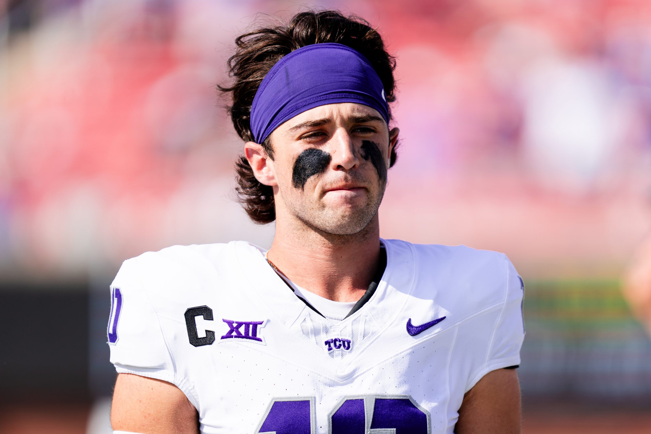 Close-up portrait of TCU quarterback Josh Hoover during a 2024 game at SMU. He wears a purple headband and white jersey, with his eyes squinted against the sun and mouth closed.