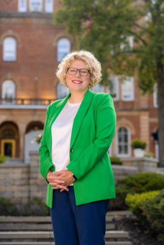 Portrait of TCU alumna and Washington & Jefferson College president Elizabeth MacLeod Walls, wearing a green blazer with a white shirt and dark pants, as she stands on a college campus on a sunny day.