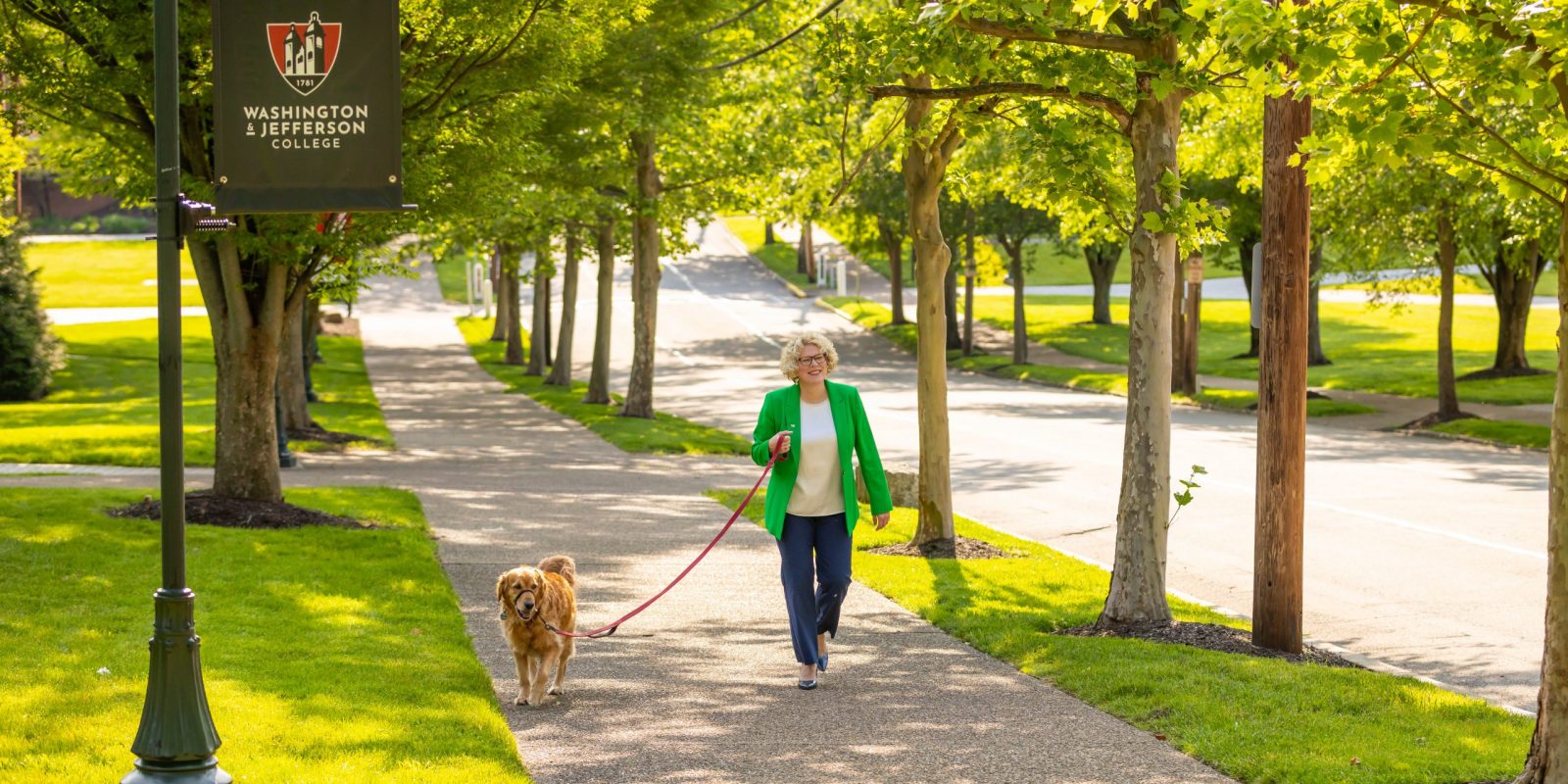 Photograph of TCU alumna and Washington & Jefferson College president Elizabeth MacLeod Walls, wearing a green blazer with a white shirt and dark pants, walking her golden retriever across a college campus on a sunny day.