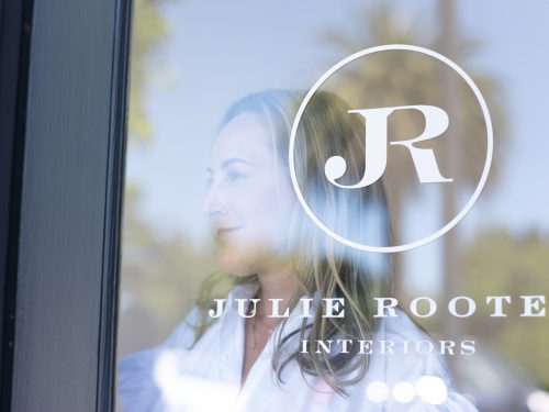 Photograph of TCU alumna Julie Rootes seen through a glass door featuring branding for Julie Rootes Interiors, with a palm tree reflected in the glass.