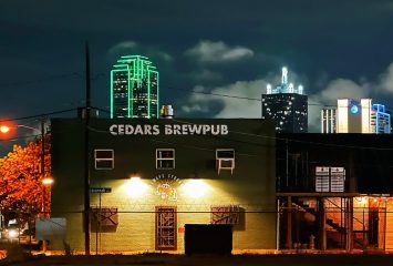 Nighttime photo of a building with 'CEDARS BREWPUB' in large letters on its wall; green- and white-lit skyscrapers rise in the background.
