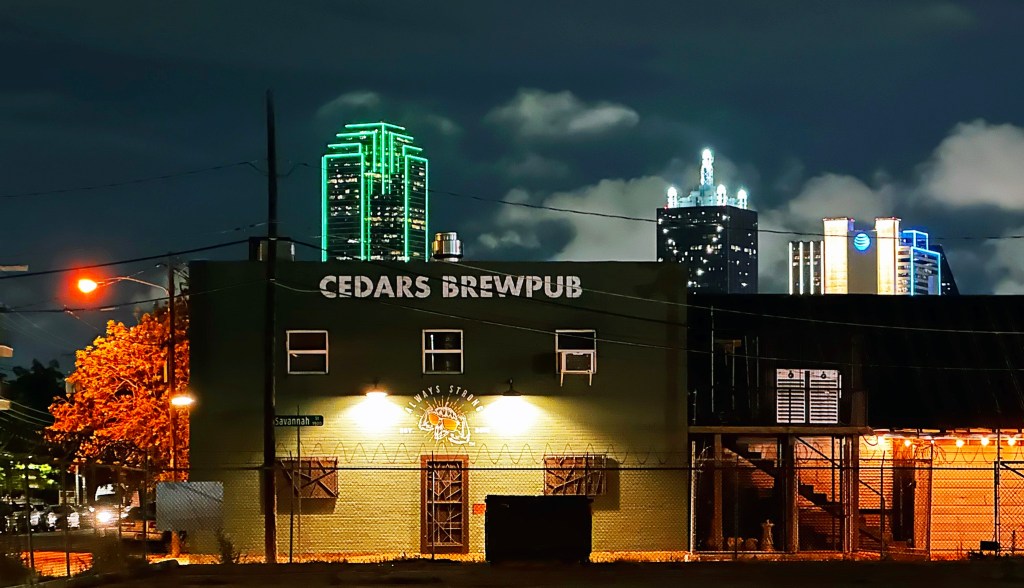 Nighttime photo of a building with 'CEDARS BREWPUB' in large letters on its wall; green- and white-lit skyscrapers rise in the background.
