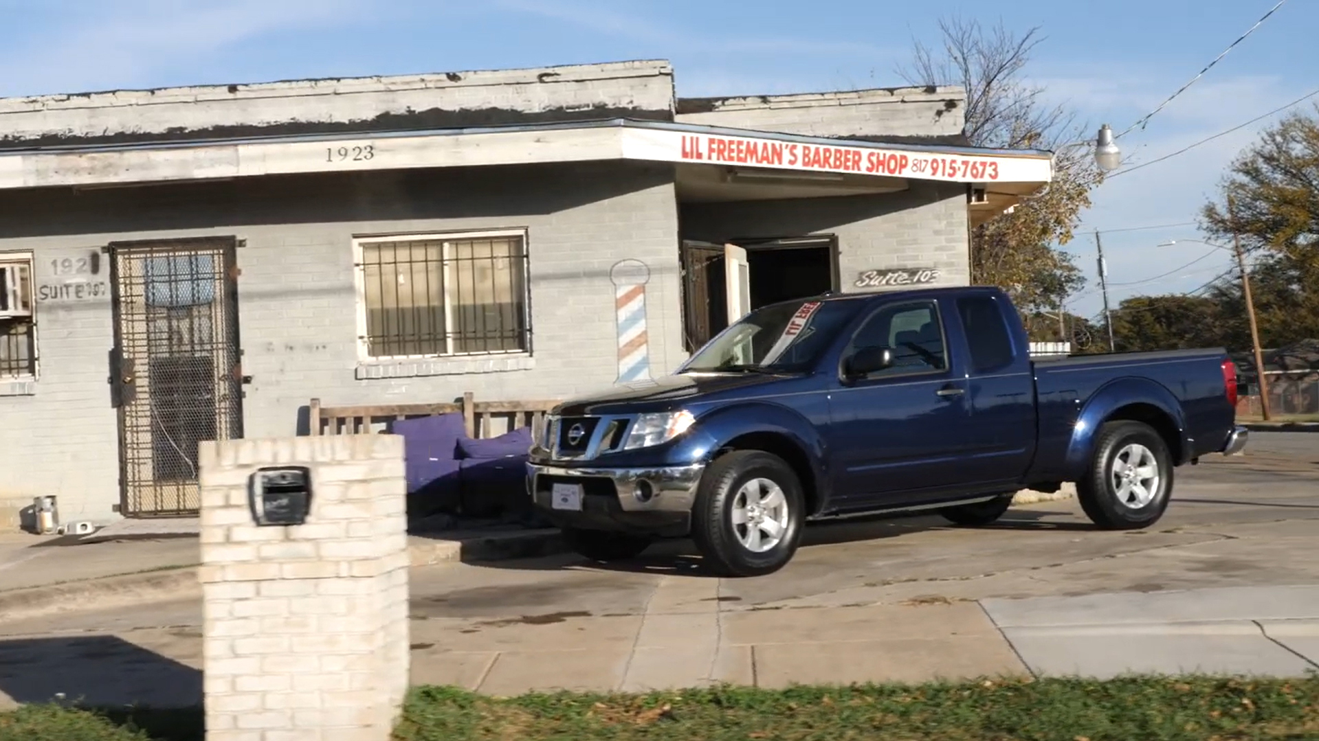 Photograph of the exterior of Lil Freeman's Barber Shop on a sunny day, with a dark blue Nissan truck parked in front.