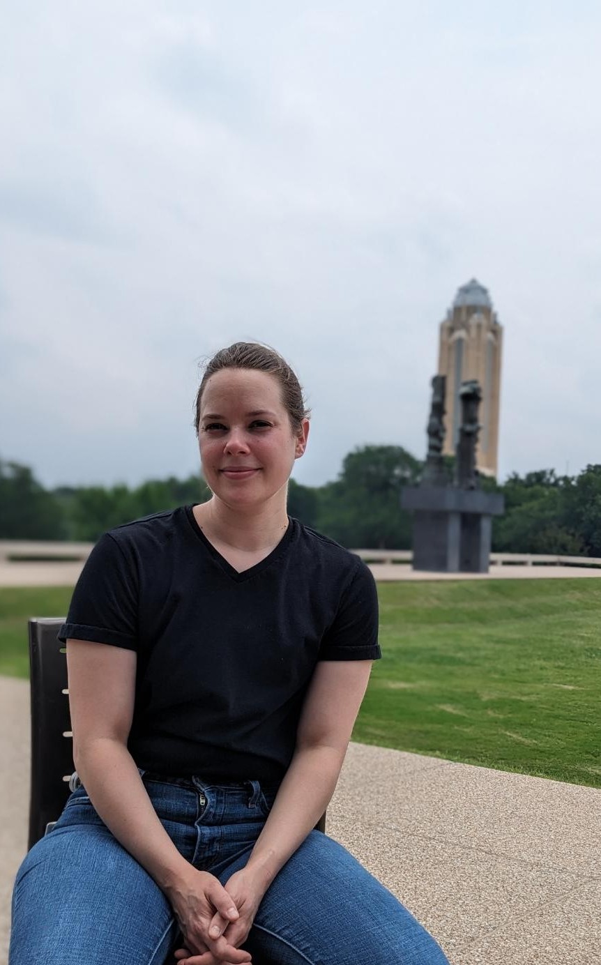 Photograph of TCU alumna Alden Williams Kresena seated outdoors, wearing jeans and a black shirt. In the background are trees, a sculpture and a tall building.