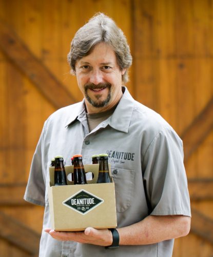 Photograph of TCU alum Dean Weaver holding a six-pack of glass beer bottles with black and white labels that read “DEANITUDE.” He stands in front of a wood-paneled backdrop, possibly a barn door.