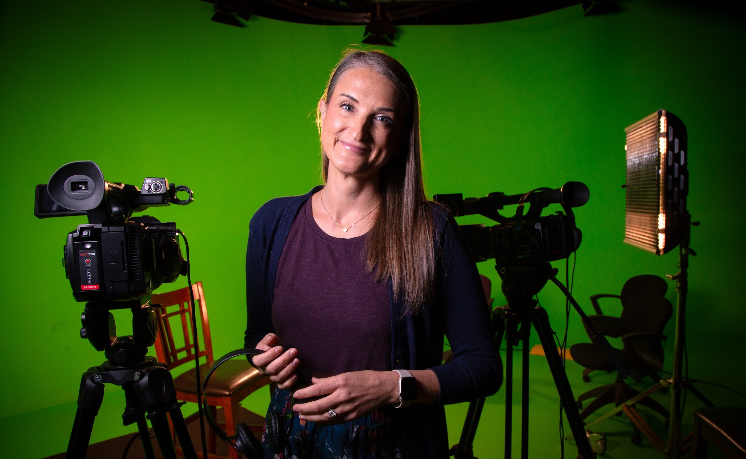 Photograph of film, television, and digital media instructor Charity Robinson standing in front of a green screen, holding a headset, with filmmaking equipment in the background.