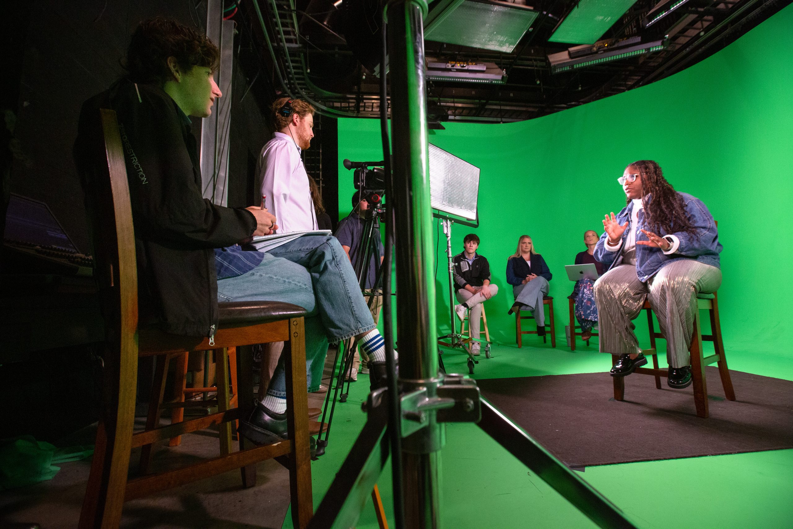 Photograph of Documentary Production students working on a project titled Trailblazers: The Create + Collaborate Story. One student sits on a stool, speaking and gesturing in front of a green screen while others observe.