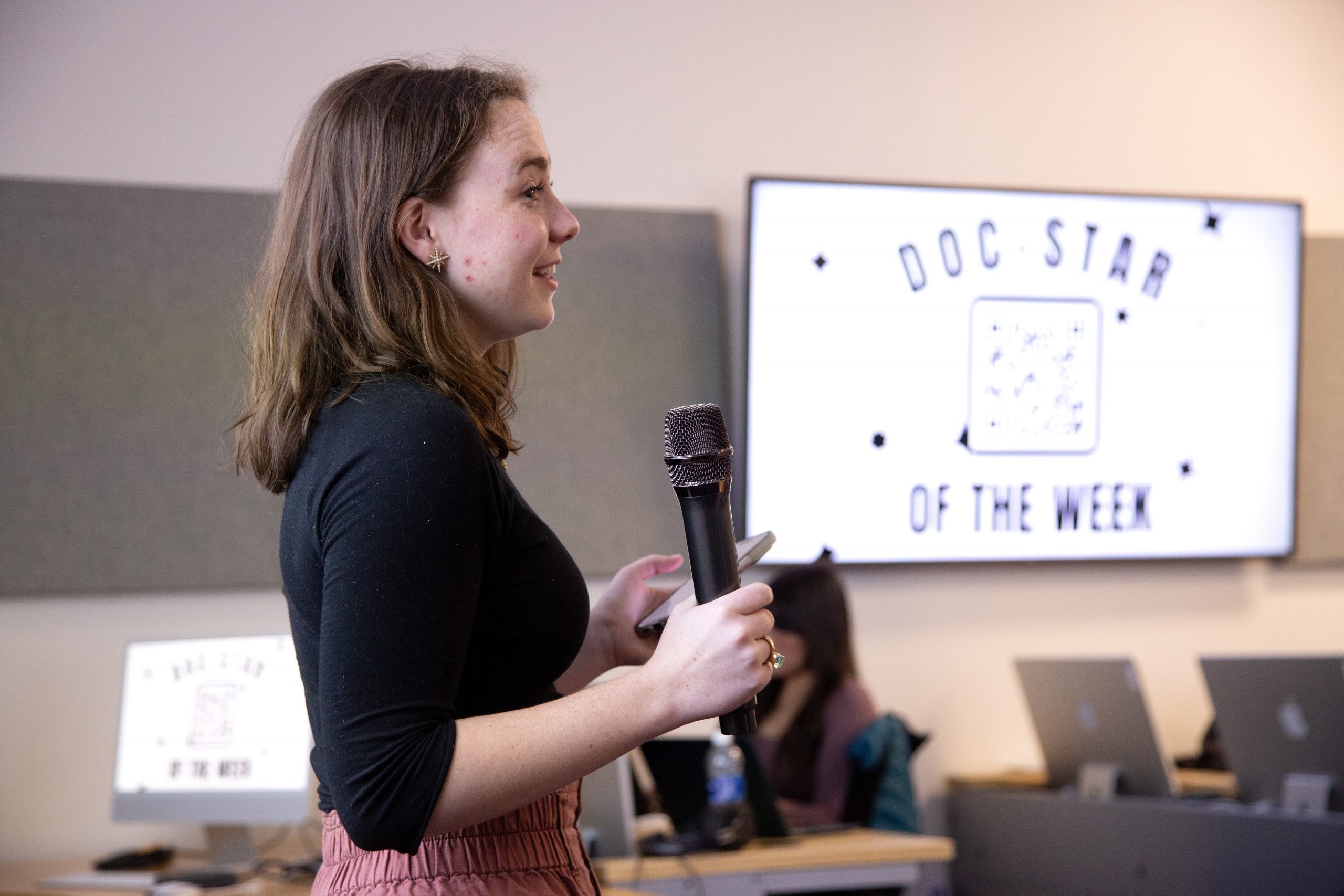 TCU student holding a microphone speaks to a class, with a board reading “Doc Star of the Week” in the background.