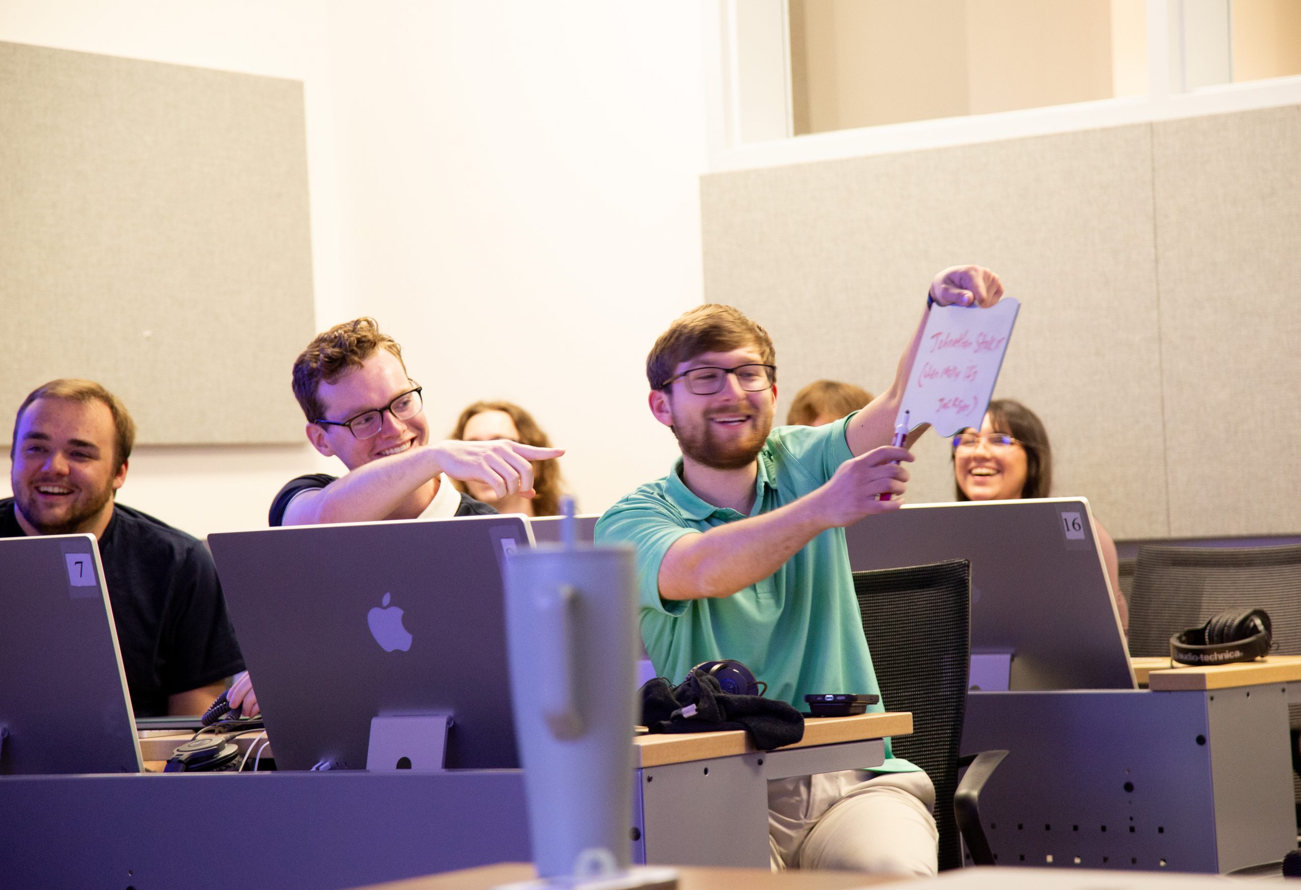 Student smiling in a classroom while holding a small whiteboard with red marker writing; other students appear to be smiling and laughing.