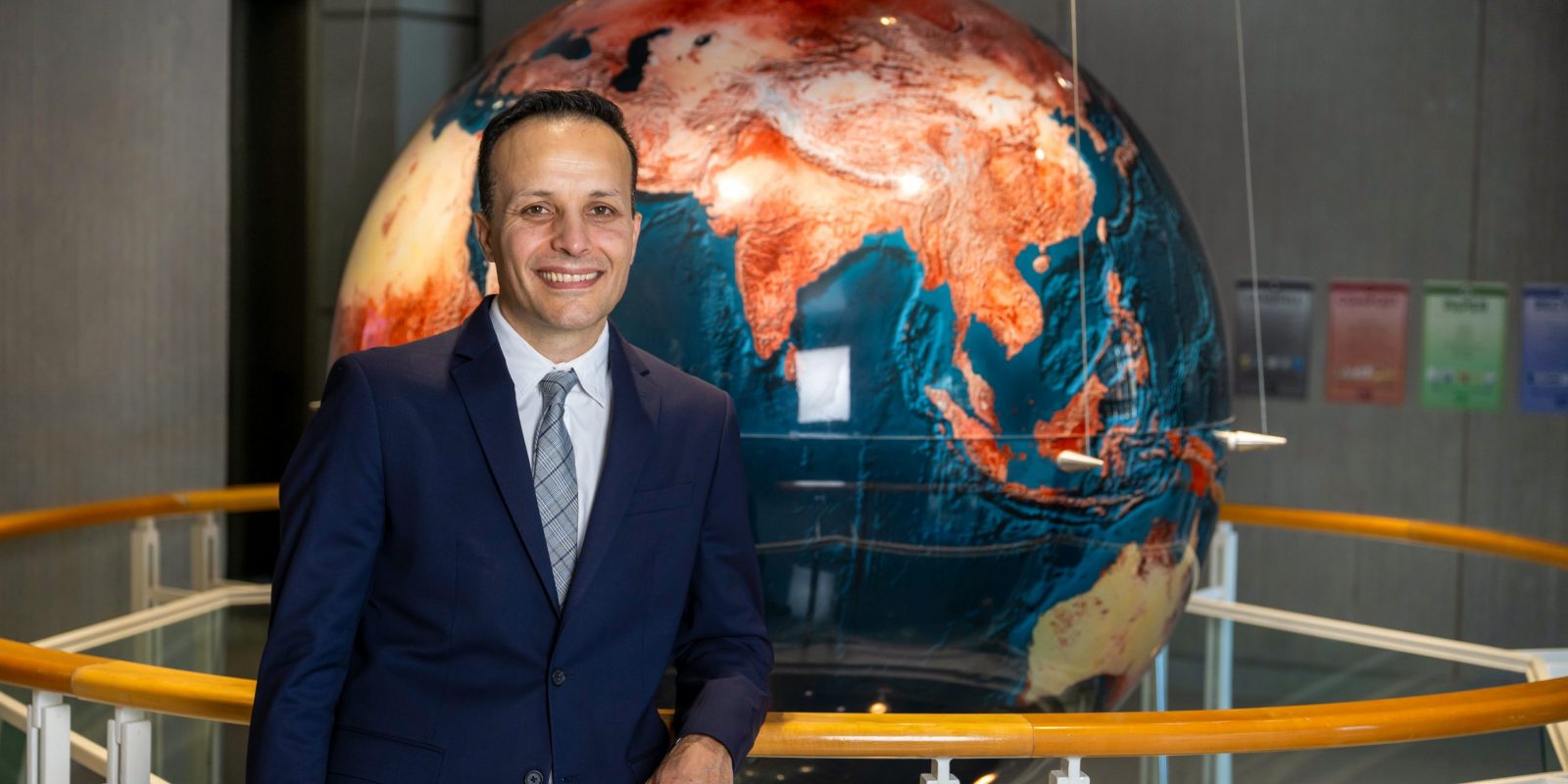 Photograph of TCU alum Alonso Sánchez in a dark suit, white shirt and gray patterned tie, leaning against a railing and smiling, with a large reddish-orange and deep blue globe behind him.