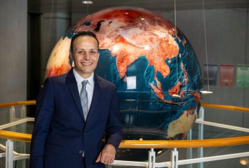 Photograph of TCU alum Alonso Sánchez in a dark suit, white shirt and gray patterned tie, leaning against a railing and smiling, with a large reddish-orange and deep blue globe behind him.