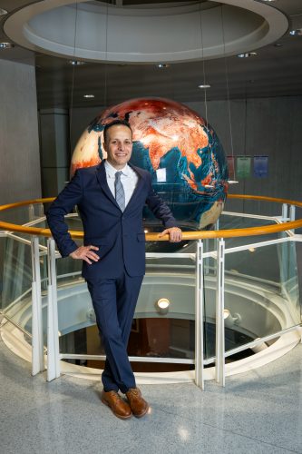 Photograph of TCU alum Alonso Sánchez in a dark suit, white shirt and gray patterned tie, leaning against a railing and smiling, with a large reddish-orange and deep blue globe behind him.