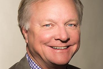 Studio portrait of TCU alumnus John Davis ’74, smiling in a purple and white checkered dress shirt and gray blazer. He is posed against a brownish-gray background.