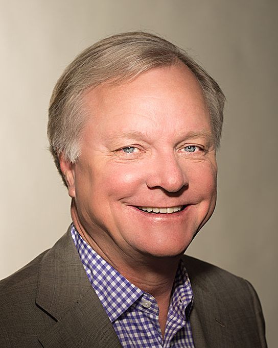 Studio portrait of TCU alumnus John Davis ’74, smiling in a purple and white checkered dress shirt and gray blazer. He is posed against a brownish-gray background.