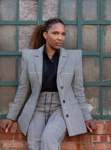 Portrait of TCU alumna Shawn Lassiter in a gray business suit, leaning against a brick windowsill with a green-framed glass window behind her.