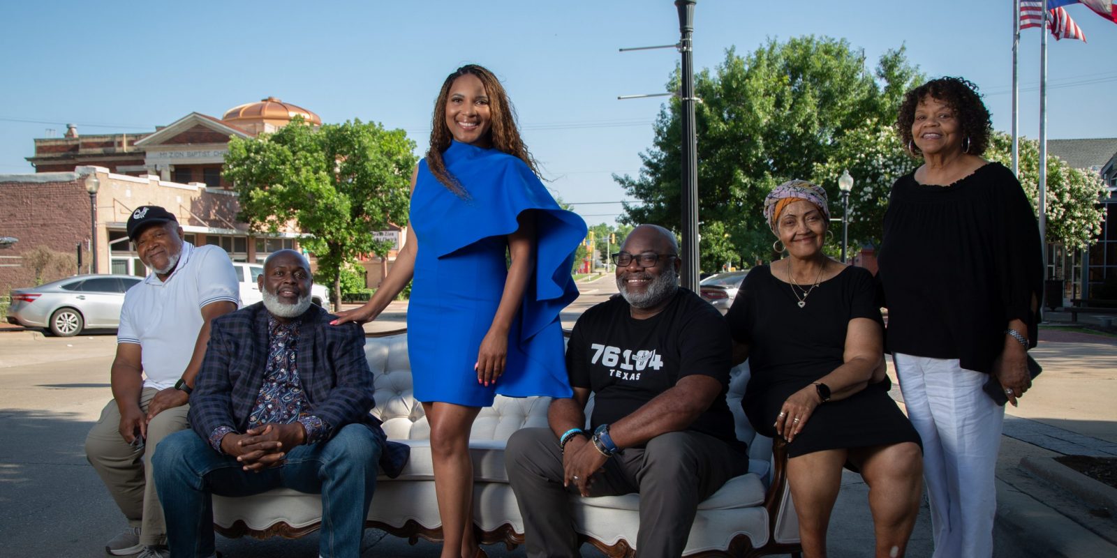 Photograph of TCU alumna Shawn Lassiter in a blue dress, standing on a street beside a white couch where five other people sit, all facing the camera. An American flag and a Texas flag appear in the top-right corner as a car drives past on a sunny day.