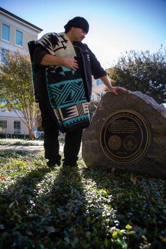 A Native American man, J. Albert Nungaray, stands next to a large stone monument with a bronze plaque. He is wearing a black beanie and a patterned blanket over his shoulders with a black jacket and pants. The blanket is black, turquoise and off-white with geometric designs. He has his right hand resting on top of the monument's smooth surface. The plaque on the monument is a bronze circle, with a statement and seals. The monument is surrounded by green plants and grass, with a building and trees visible in the background. The sun is shining brightly, casting shadows on the ground.
