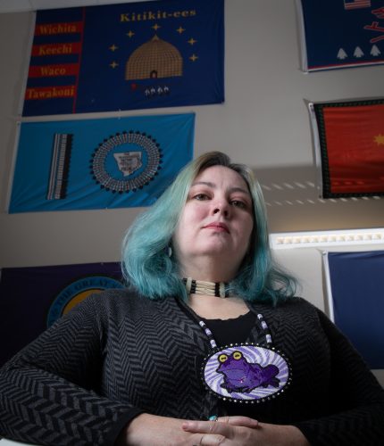 A portrait of a woman with teal-streaked hair, a dark sweater and a black shirt. She wears a multi-strand necklace with white beads and a large, beaded pendant featuring a purple frog with yellow eyes and a turquoise bead at the bottom. She is looking directly at the camera with a neutral expression. Behind her, several tribal flags are visible, including one with the names of the Wichita, Keechi, Waco and Tawakoni tribes and another with a circular design in blue.