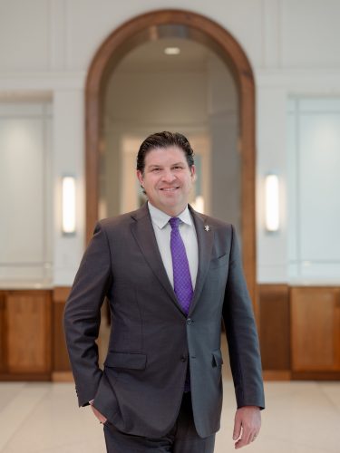 Photograph of TCU Chancellor Daniel Pullin, wearing a purple tie, dress shirt and dark grey suit, standing in a hallway of the Harrison building on campus.