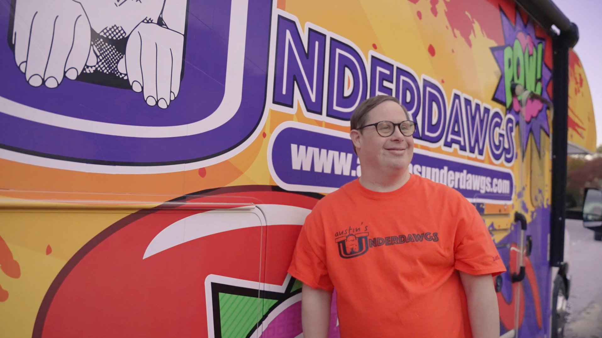 Photograph of Austin Underwood, a man with Down syndrome, standing beside a yellow-orange hot dog truck with the word "UNDERDAWGS." He wears black-framed glasses and an orange t-shirt.