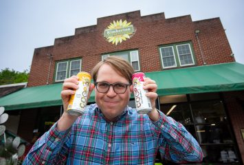 Photograph of TCU alum Cooper Neel, wearing black-framed glasses and a blue plaid button-down shirt, smiling while holding cans of "Coop" beverages in front of a brick building with a green overhang. A yellow, green and orange sunflower emblem on the building reads "Kimberton Whole Foods."