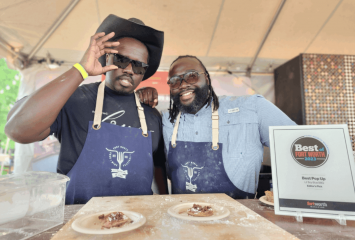 Photograph of brothers Reggie (left) and Cedric Robinson standing behind a table with two plates of food. Both are smiling — Reggie holds his right hand to his sunglasses, while Cedric leans slightly toward his brother.