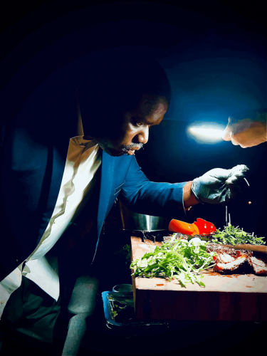 Photograph of Reggie Robinson standing in a dark space wearing a sport jacket, dress shirt and culinary gloves. He inspects a slab of meat on a table illuminated by a flashlight, alongside pieces of lettuce and tomato.