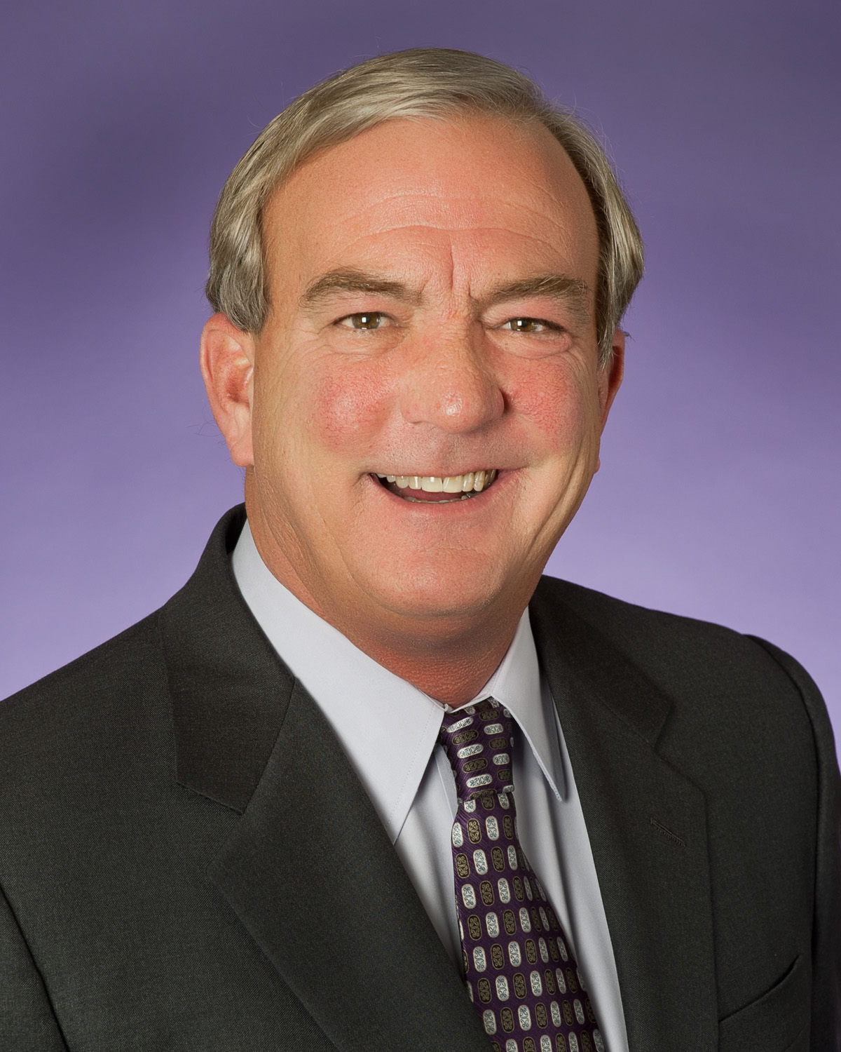 Portrait of TCU alum Clarence Scharbauer III (Class of 1973) wearing a black sport coat, dress shirt and patterned tie, smiling as he poses against a grayish-purple background.