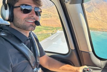 Photograph of TCU student Matthew Miamidian piloting an aircraft, wearing sunglasses and an aviation headset, with a view of water and land visible through the cockpit windows.
