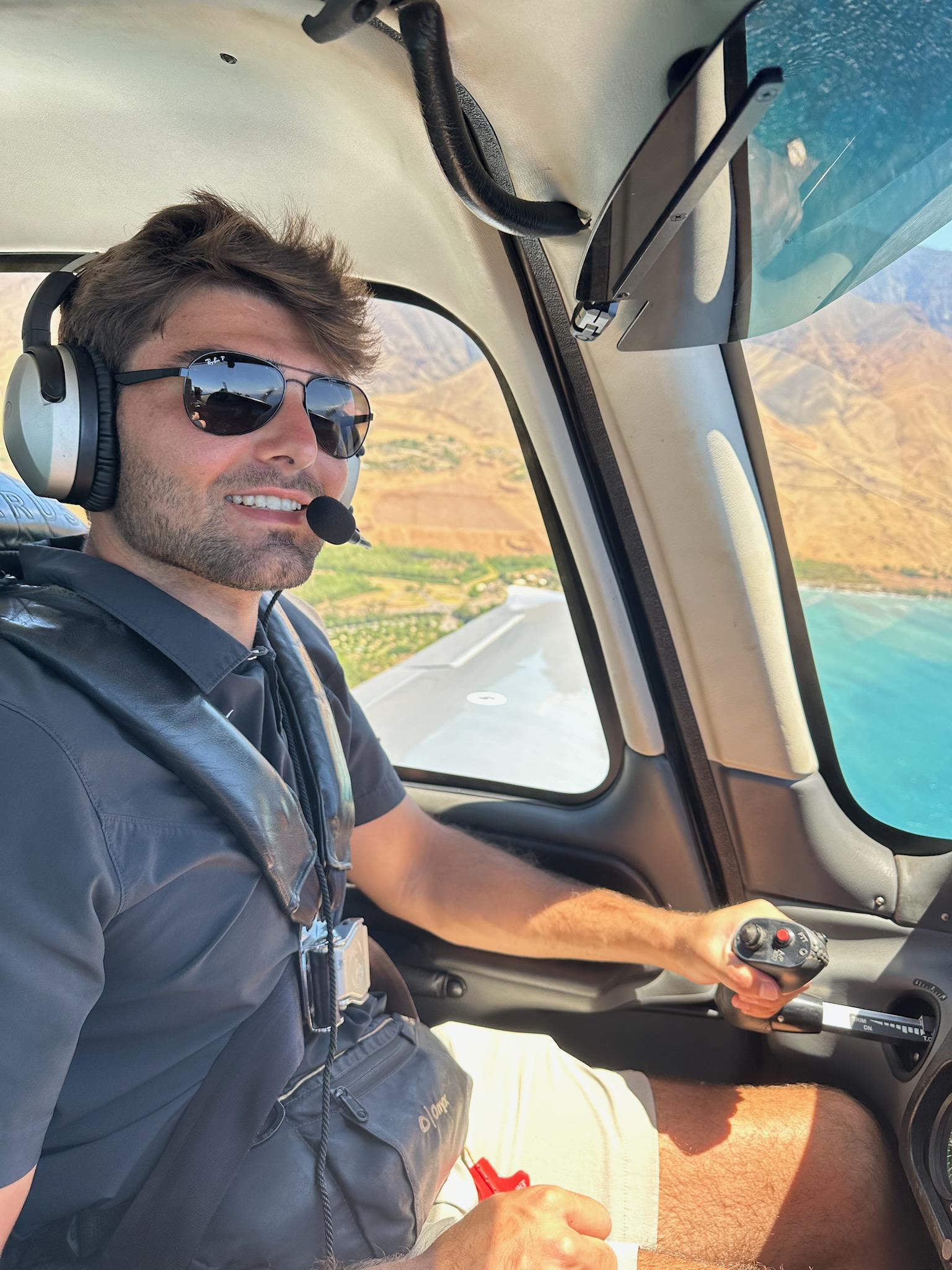 Photograph of TCU student Matthew Miamidian piloting an aircraft, wearing sunglasses and an aviation headset, with a view of water and land visible through the cockpit windows.