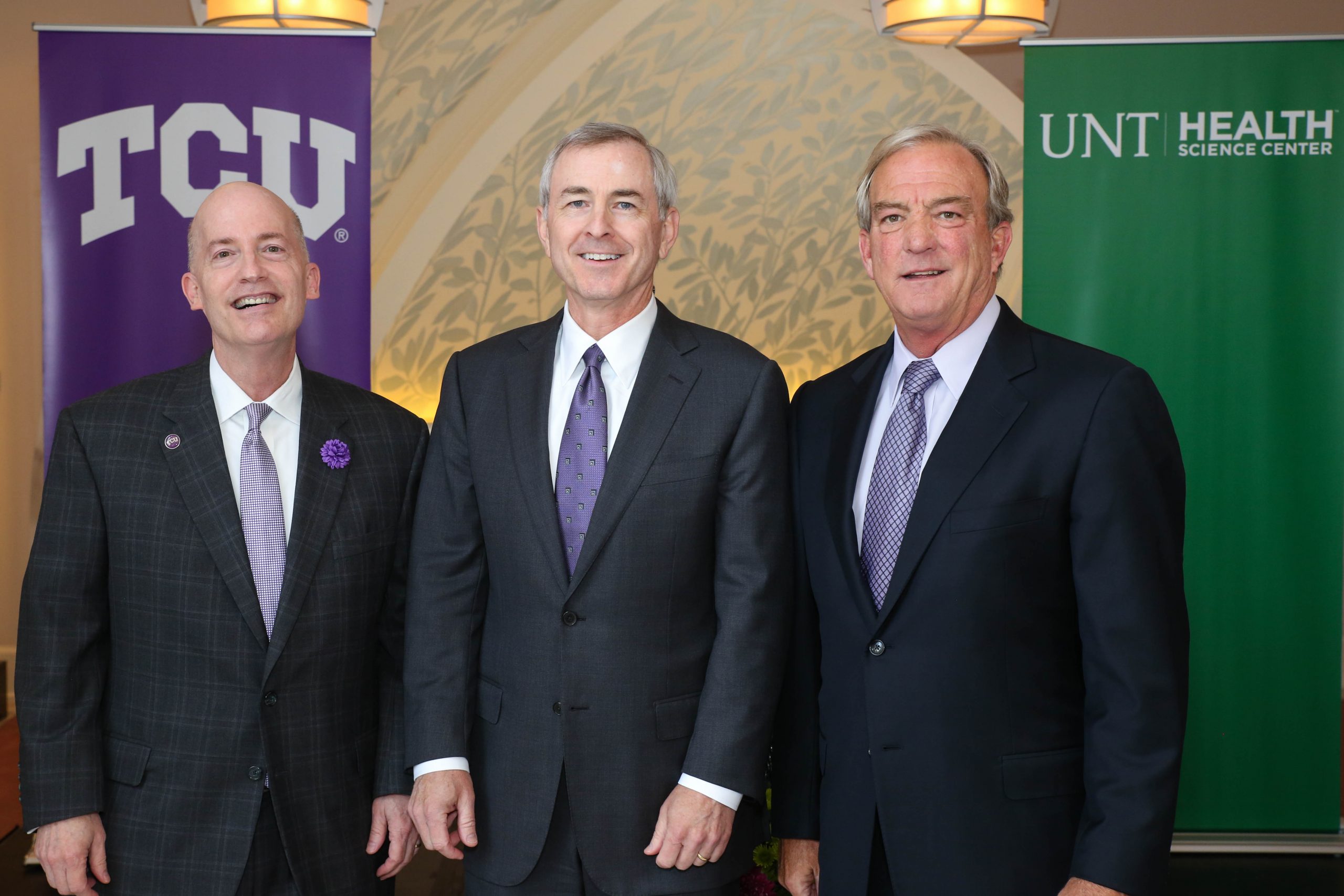 Photograph of Clarence Scharbauer III, Mark Johnson and Victor J. Boschini, Jr., dressed in formal attire and standing in front of “TCU” and “UNT Health” promotional signs.