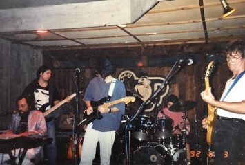 Photograph of five musicians performing on a stage, with a tan and black sign reading “The Hop” pinned to the wood-paneled wall behind them. The group plays guitar, bass, keyboards and drums.