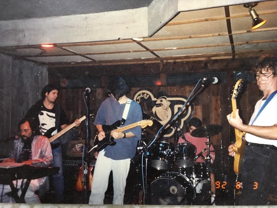 Photograph of five musicians performing on a stage, with a tan and black sign reading “The Hop” pinned to the wood-paneled wall behind them. The group plays guitar, bass, keyboards and drums.