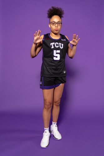 Photograph of Horned Frog student-athlete Olivia Miles wearing a black TCU basketball uniform against an all-purple backdrop. She holds the jersey forward, hands around the "TCU" emblem on the front.
